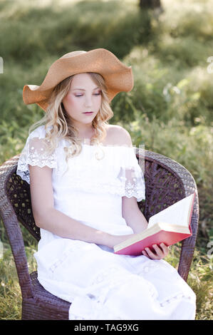 Belle fille blonde 15-16 ans reading book sitting in chair à l'extérieur. Banque D'Images