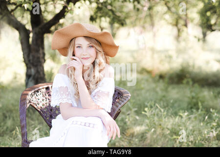 Belle fille blonde wearing hat et white dress sitting in chair à l'extérieur. Banque D'Images