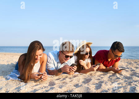 Groupe de millénaire à l'aide portant sur les smartphones ensemble serviette de plage près de la mer sur le coucher du soleil d'été. Les jeunes personnes intoxiquées par les téléphones mobiles intelligents. Toujours Banque D'Images