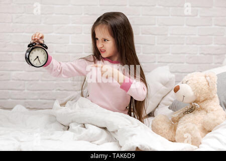 En colère petit enfant fille en pyjama avec réveil au lit. Bon matin Banque D'Images
