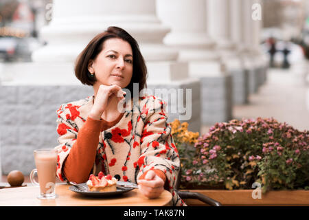 Femme élégante 40-45 ans manger un gâteau et de boire du café dans le café en plein air. En regardant la caméra. Banque D'Images
