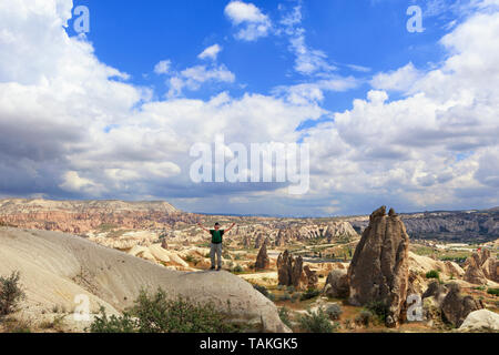 Un jeune homme en T-shirt vert et sac à dos sur son dos est debout au sommet d'une colline en Cappadoce et à la recherche jusqu'à la blue cloudy sky avec ses bras Banque D'Images