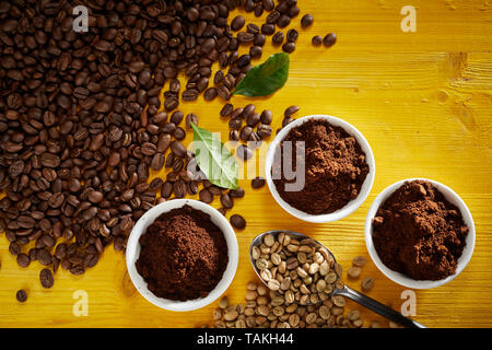 Les grains de café torréfié et de matières premières avec de nouveaux motifs dans des petits bols vue d'en haut sur bois jaune avec deux feuilles vertes et copy space Banque D'Images