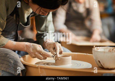 Close-up of Asian artisane dans apron assis à la poterie et à l'aide de l'outil d'artisanat de roue tout en façonnant l'argile humide navire Banque D'Images