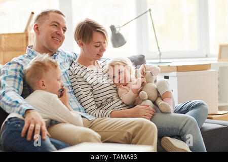 Rêve sourire jeune famille de tenues décontractées assis sur un grand canapé et enlacés tout en passant du temps ensemble à la maison Banque D'Images