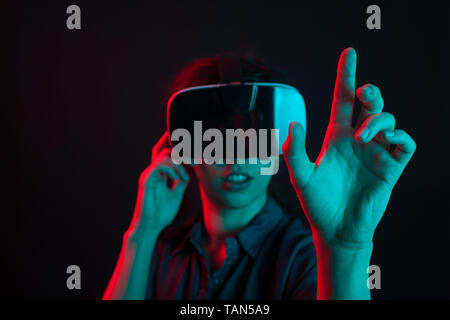 Belle jeune femme gaie divertis avec casque de réalité virtuelle en studio et éclairage coloré. Jeune femme à l'aide de la technologie moderne. Femme avec casque de réalité virtuelle de toucher l'air. Banque D'Images