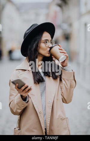 Jeune femme avec du café pour aller debout à la rue et à l'aide de mobile phone Banque D'Images