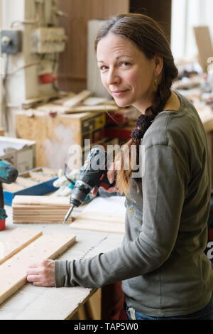 Jeune femme à l'aide d'un tournevis électrique sur un morceau de bois à l'atelier Banque D'Images