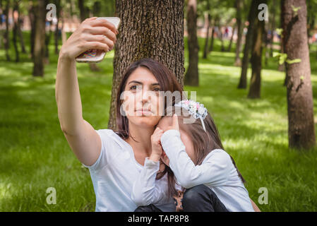 Jeune maman et petite fille prend ensemble selfies.petite fille cache son visage.Photo de jeune mère et sa fille ayant sur l'herbe verte selfies Banque D'Images