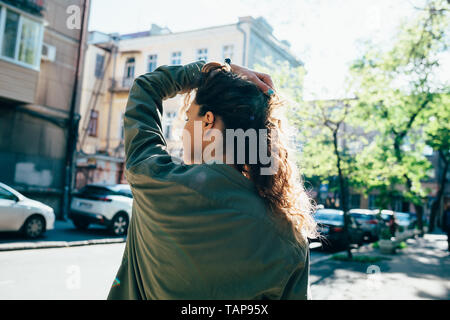 Vue arrière du jeune femme portant chemise verte debout sur la rue en soulevant les cheveux avec sa main. Banque D'Images