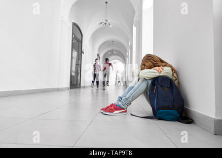 Fille avec de longs cheveux en shirt, jeans et baskets appuyée sur les genoux. Seul, triste lycéenne assis sur sol gris avec de courber la tête. Sac à dos enfant shool près de. Banque D'Images