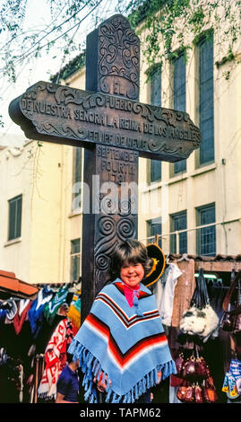 Une jeune fille dans un souvenir sarape pose devant une énorme croix en bois qui commémore la fondation de la 1781 El Pueblo de Los Angeles, maintenant la ville de Los Angeles, Californie, USA. L'inscription en espagnol, El Pueblo de Nuestra Señora la Reina de los Ángeles, traduit comme la ville de Notre-Dame la Reine des Anges. Est également inscrit le nom de la ville fondateur et Gouverneur de Californie espagnole, Felipe de Neve. La croix se dresse à l'extrémité sud de Olvera Street, qui a été un marché mexicain depuis 1930 et attire les visiteurs avec des stands de fournisseurs, magasins et restaurants. Banque D'Images