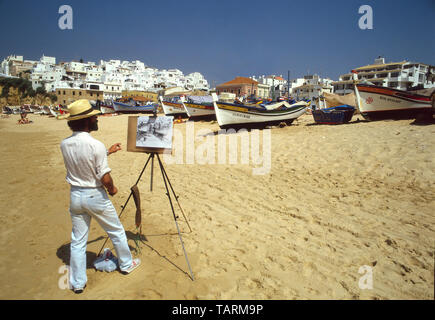 Man1980s artiste au travail sous ciel bleu clair avec tableau blanc esquisse sur bateau de pêche et la plage de sable propre de soleil vacances Albufeira Algarve Portugal Banque D'Images