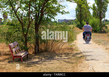 Un banc à l'ombre d'un arbre, vieux et patiné, dans le contexte d'un paysage rural et d'un résident qui est de la trottinette le long d'une vieille concr Banque D'Images