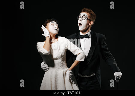 Portrait de couple surpris mime sur fond noir. man in tuxedo et verres et femme en robe blanche Banque D'Images