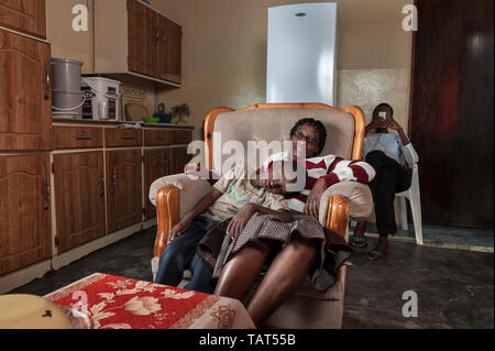 Mère dans un fauteuil tenant son jeune sone comme il se repose, fils aîné dans l'arrière-plan avec cell phone Banque D'Images