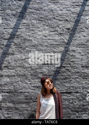 Belle à la mode hipster asian woman wearing white sleeveless, lunettes et motif à pois rouge turban smiling sur mur de brique gris moderne retour Banque D'Images