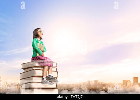 Jolie fille asiatique avec des lunettes tenant le livre tout en restant assis sur la pile de livres avec ville et fond de ciel bleu. Concept Retour à l'école Banque D'Images