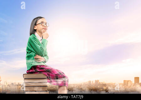 Jolie fille asiatique avec des lunettes pense que tout en étant assis sur la pile de livres avec ville et fond de ciel bleu. Concept Retour à l'école Banque D'Images