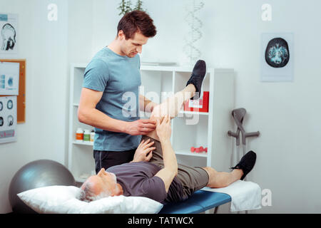 La thérapie professionnelle. Bel homme du métier, debout près de son patient tout en effectuant un massage de jambe pour lui Banque D'Images