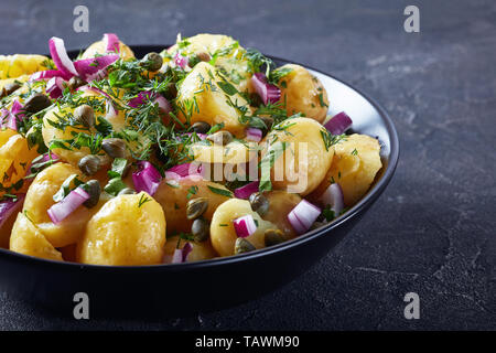 Close-up of Savoury allemand nouveau salade de pommes de terre avec l'oignon rouge, les câpres, les verts et la moutarde de vinaigre dans un bol noir sur une table, la vue à partir de Banque D'Images