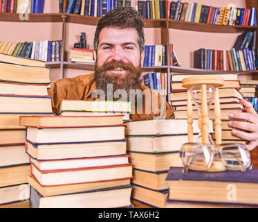 Concept bibliothécaire. Enseignant ou étudiant avec barbe est assis à table avec des livres, des flous artistiques. Sur l'homme heureux face entre les piles de livres, tout en étudiant en bibliothèque, des étagères sur l'arrière-plan. Banque D'Images