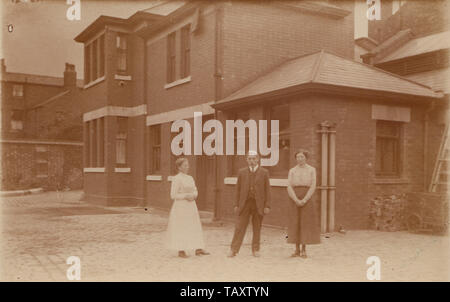 Vintage Carte postale photographique montrant deux femmes et un homme se tenait sur une rue pavée à l'extérieur d'un grand bâtiment. Peut-être un lieu de travail. Banque D'Images