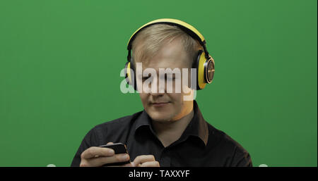 Guy écoute de la musique sans fil casque jaune en. Portrait of attractive man using smart phone pour écouter la musique dans la prise casque tout en étant assis près de l'écran vert. Chroma key Banque D'Images