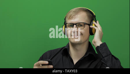 Guy écoute de la musique sans fil casque jaune en. Portrait of attractive man using smart phone pour écouter la musique dans la prise casque tout en étant assis près de l'écran vert. Chroma key Banque D'Images