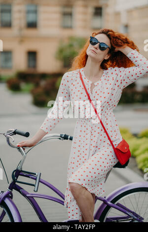 Activité de plein air concept. Belle femme aux cheveux rouges bouclés avec expression rêveuse, utilise location pour voyager à l'échelle de la ville, à la mode porte whit Banque D'Images