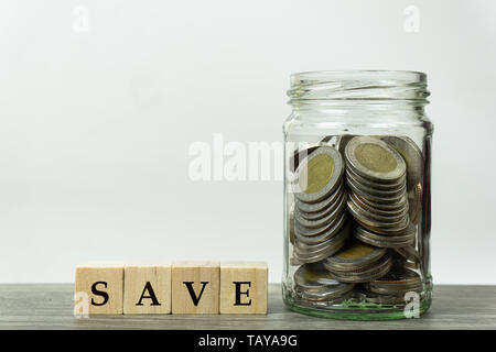 Concepts d'économiser de l'argent. Pièces empilées dans un bocal en verre et la cale en bois sur la table en bois avec un fond blanc. Banque D'Images