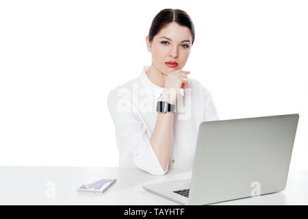 Portrait de femme brune succès graves en chemise blanche assise avec un ordinateur portable, touhing son menton et à la recherche à l'appareil photo avec visage pensif. Banque D'Images