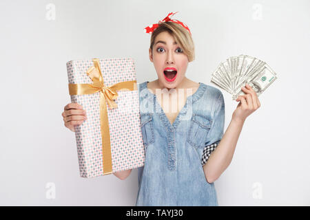 Pas moyen ! Portrait de jeune fille en étonné chemise en jean décontracté avec un miroir et bandeau rouge debout, montrant boîte cadeau et dollars de ventilateur avec bouche ouverte. Banque D'Images