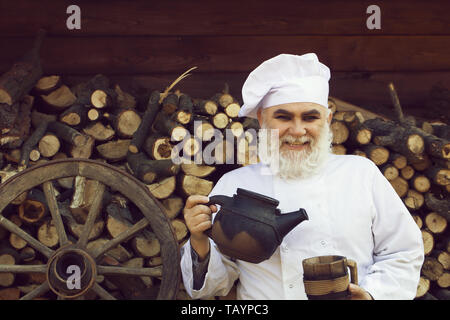 Chef cuisinier homme barbu en uniforme et chapeau à longue barbe sur le visage souriant de vieux fer holding plateau électrique et coupe du bois près de roue sur fond de bois. Banque D'Images