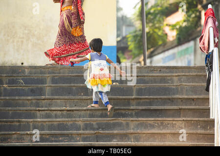 Une petite fille portant une robe colorée est de monter les escaliers derrière sa mère qui s'habille le sari, une robe traditionnelle indienne. Varanasi, Inde. Banque D'Images