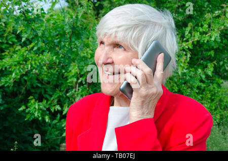 Portrait de vieille dame, heureuse de parler, parler. sourire grand-mère répondant à un appel à sun day. moderne caucasian granny en veste rouge attente téléphone près de il Banque D'Images