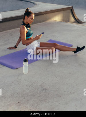 Jeune femme dans les vêtements de sport assis sur les tapis d'exercice et à l'aide de son téléphone intelligent. Contrôle de performance physique sa femme sur téléphone mobile après l'entraînement. Banque D'Images