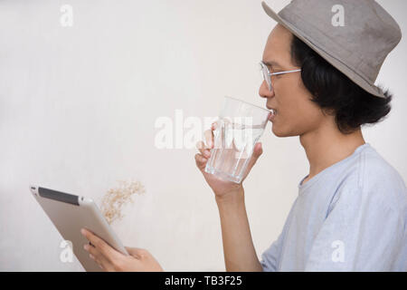 Young Asian man avec chapeau et lunettes surpris par une tablette ou un ordinateur portable alors que boire un verre d'eau Banque D'Images