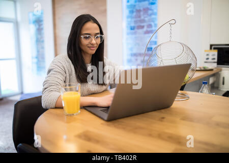 Beautiful woman relaxing with her laptop tout en tenant un verre de jus d'orange dans la cuisine Banque D'Images