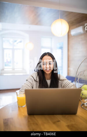 Beautiful woman relaxing with her laptop tout en tenant un verre de jus d'orange dans la cuisine Banque D'Images