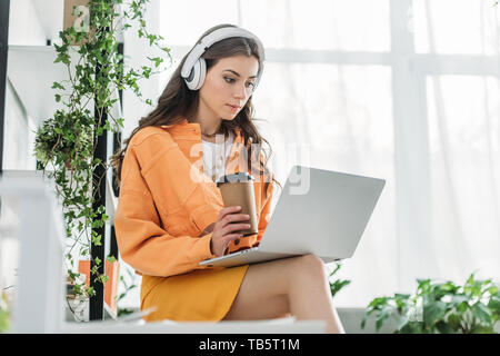 Jeune femme attentive holding Coffee cup lors de l'utilisation d'ordinateur portable et l'écoute au casque de la musique Banque D'Images