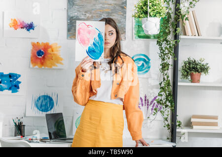 Belle jeune fille se cacher derrière la peinture tout en se tenant dans cette chambre spacieuse est décorée avec des plantes vertes et des dessins sur mur blanc Banque D'Images