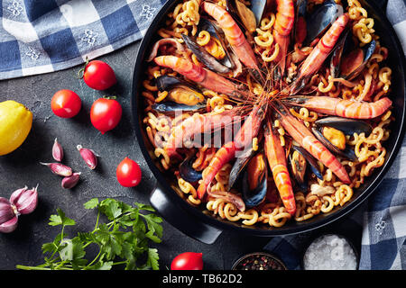 Une paella, Fideua espagnole comme plat à pâtes spéciales grosses crevettes, chair de poisson blanc, les calmars, les moules dans une casserole noire sur une table en béton avec ingredi Banque D'Images