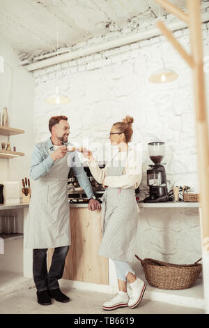 Lunettes qui retentit. Deux baristas joyeuse portant des tabliers debout dans la cuisine et boire le café ensemble. Banque D'Images