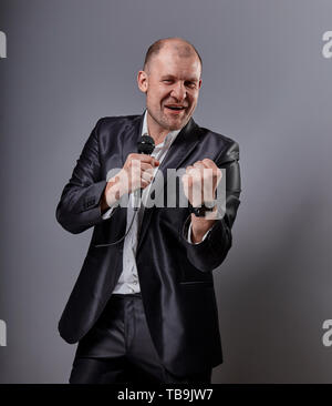 Déménagement émotionnel sourire heureux homme artiste présentant le spectacle holding micro à la main et montrant le succès signe le poing sur zone de couleur gris Banque D'Images