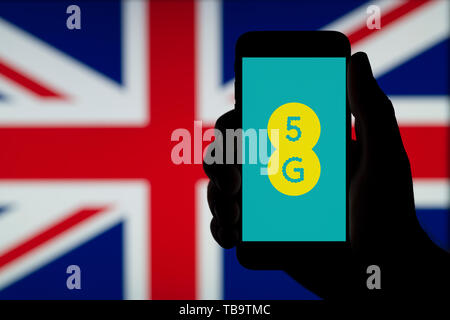La silhouette d'une main d'un homme est titulaire d'un smartphone affichant le logo de l'ERE 5G, devant un drapeau britannique (usage éditorial uniquement). Banque D'Images