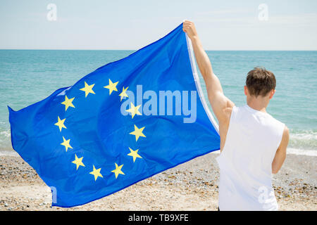 Jeune adulte caucasien homme tenant sur une plage, tenant le drapeau de l'Union européenne Banque D'Images