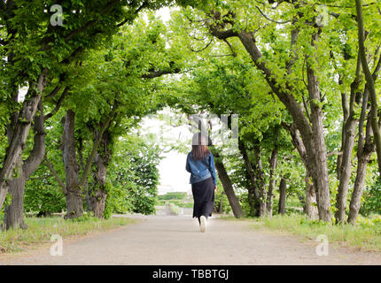 Young Asian woman walking in a green alley avec des arbres. Route qui traverse un parc au printemps. Concept de voyage. Banque D'Images