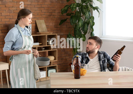 Lazy mari boire de la bière pendant que sa femme à la cuisson des aliments Banque D'Images