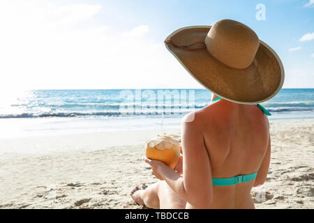 Vue arrière d'une Woman in Bikini Hat Sitting on Sand Holding Coconut At Beach Banque D'Images